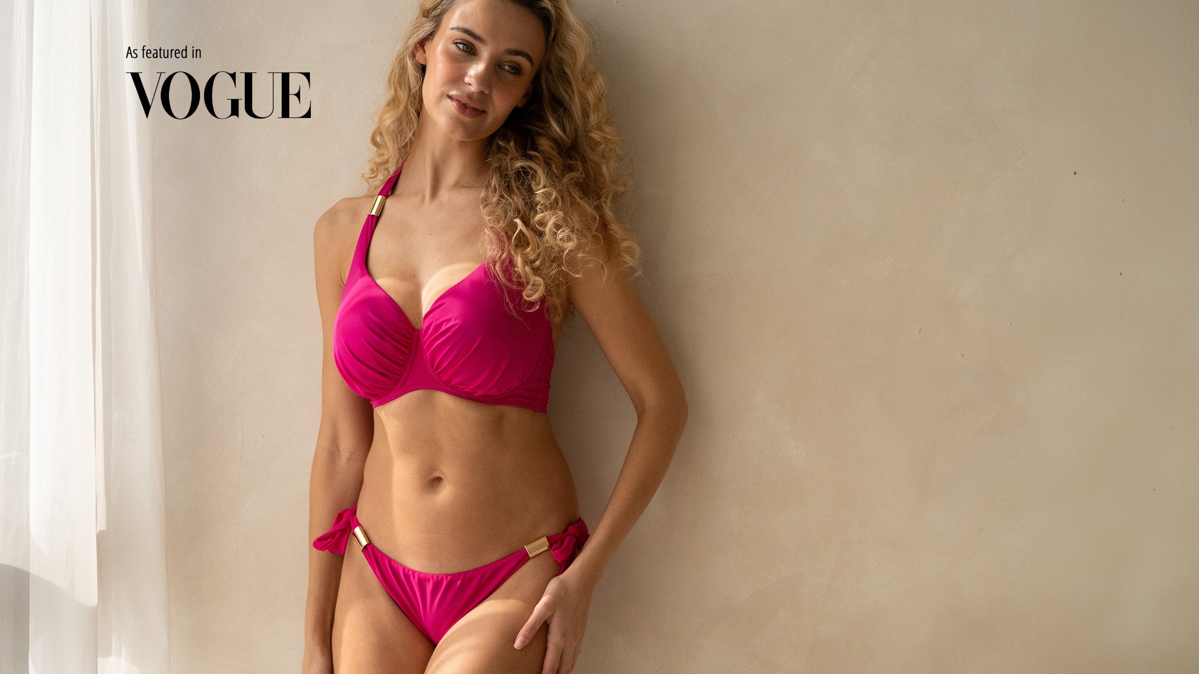 Woman in a pink halter neck bikini for fuller busts standing against a beige wall with 'Vogue' branding.